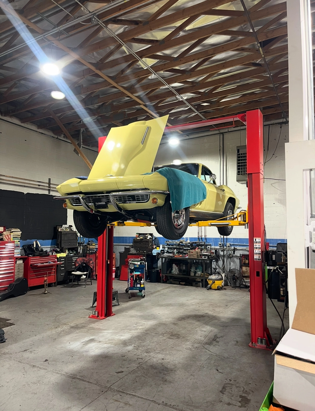 Yellow classic Corvette raised on a hydraulic lift with the hood open inside an auto restoration workshop.