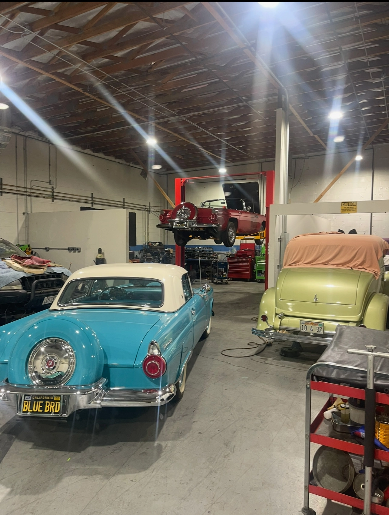 Multiple vintage cars inside a classic auto restoration workshop, including a blue classic car and several vehicles in various stages of repair.