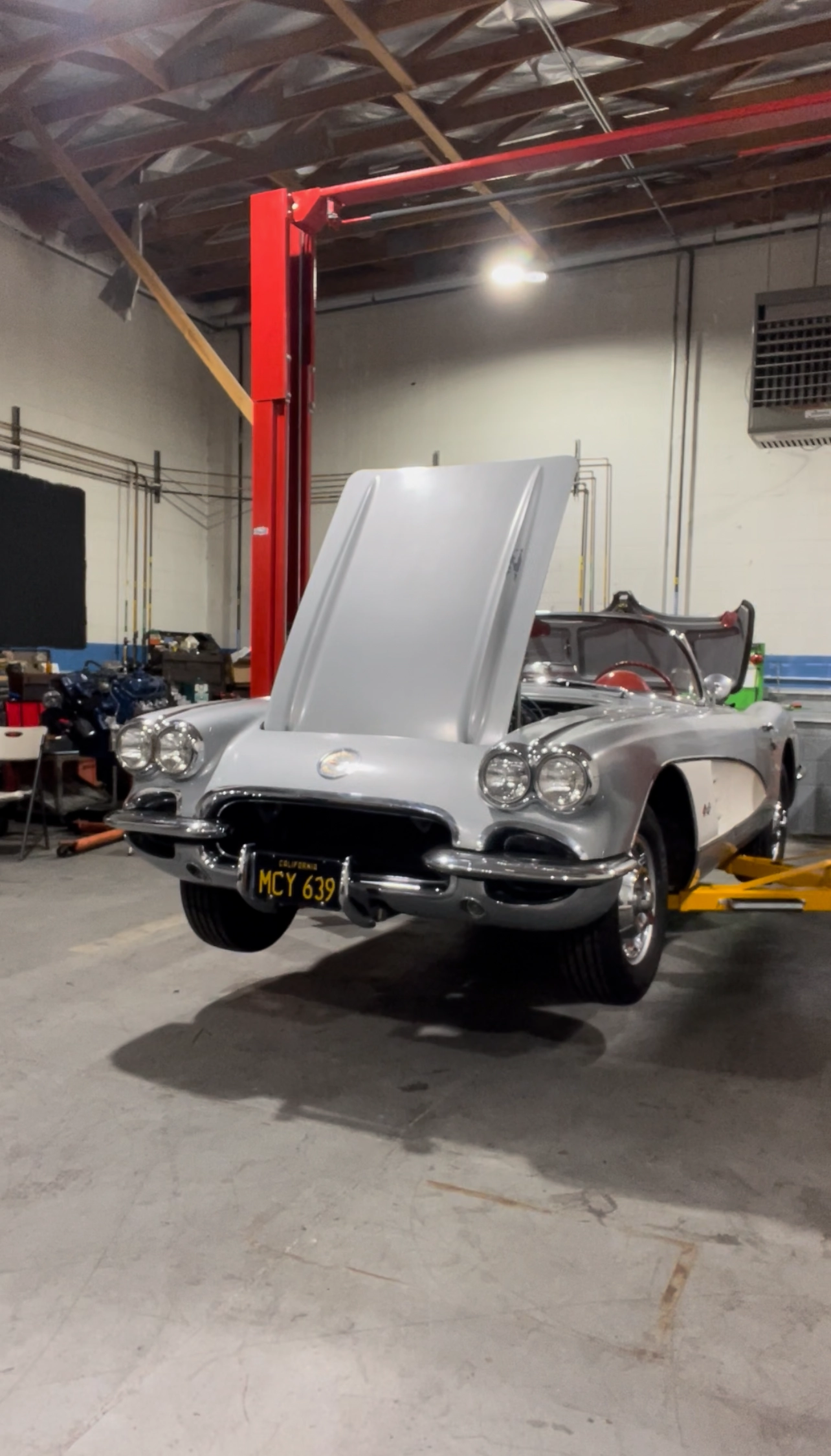 Silver classic Corvette convertible on a lift with the hood open inside an auto restoration workshop.